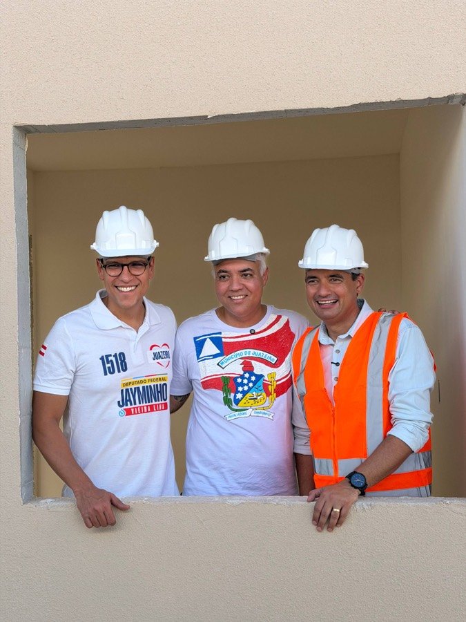 Jayminho de capacete ao lado de dois homens na janela de uma casa em construção